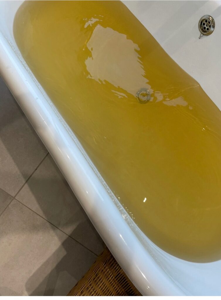 Interior scene showing a domestic bathtub filled with discoloured water, appearing yellow to brown in colour. The bathtub is white enamel and partially filled, with the water exhibiting a cloudy and opaque quality rather than clear appearance. The discolouration is uniform across the bath, with a slightly lighter patch visible where water movement or reflection occurs near the centre. A metal drain fitting is visible at the end of the bath, and surrounding surfaces appear clean and intact. The colour and clarity of the water indicate a significant deterioration in aesthetic water quality, likely associated with elevated turbidity, iron or manganese presence, or disturbance within the distribution system. This condition would be unacceptable for normal consumption and use, and may result in consumer complaints relating to discolouration, taste, or confidence in the supply. The image illustrates a clear example of discoloured water reaching a consumer property, highlighting the importance of maintaining distribution system integrity and effective control of particulates within the network.