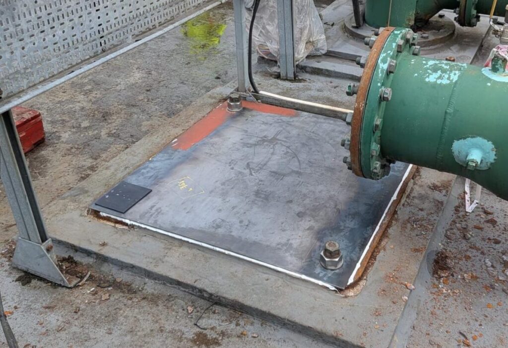 Exterior view of a water supply installation following remedial works, showing a previously exposed access point now securely sealed with a fitted metal cover. The cover is fixed in place with large bolts at each corner and sealed around the perimeter, forming a continuous barrier between the access chamber and the surrounding environment. The plate sits flush within a concrete surround, reducing the potential for gaps or ingress pathways. Adjacent pipework remains visible, including large diameter green pipes with flanged connections, but the immediate area around the access point is now clear of loose materials and temporary coverings. Cables and equipment appear tidied and better managed compared to the previous condition. The surrounding hardstanding is intact, and the area is contained within a structured framework, with no reliance on temporary barriers. The completed arrangement demonstrates improved asset security and protection against contamination ingress, with the fixed, sealed cover providing a robust and permanent solution. The removal of loose materials and proper enclosure of the access point reduces both hygiene risks and physical hazards, supporting compliance with expectations for safeguarding drinking water infrastructure.