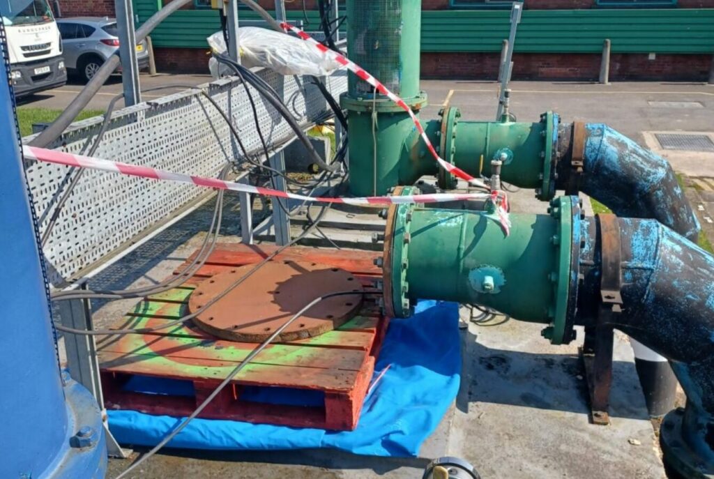 Exterior view of a water supply installation area showing exposed pipework and fittings associated with distribution infrastructure. Large diameter green pipes with flanged connections are mounted above ground, with visible sections of older, corroded blue pipework. Adjacent to the pipework is an open or uncovered access chamber, temporarily sealed with a circular metal plate placed on a wooden pallet rather than being fixed in position. Electrical cables and equipment are present and loosely arranged across the area, with some cabling draped over the structure. A temporary barrier using red and white hazard tape is positioned around the installation, indicating restricted access, but the containment appears informal and not fully secured. A blue tarpaulin sheet is placed beneath part of the arrangement, possibly to protect the surface or collect debris. The surrounding area includes hardstanding and nearby buildings, with vehicles parked in the background. The overall condition suggests ongoing works or maintenance activity; however, the exposed access point, unsecured cover, and presence of loose materials may present risks of contamination ingress, physical hazards, or unauthorised access if not adequately controlled. The arrangement highlights the importance of secure sealing of infrastructure, proper site safeguarding, and management of potential contamination pathways within drinking water systems.