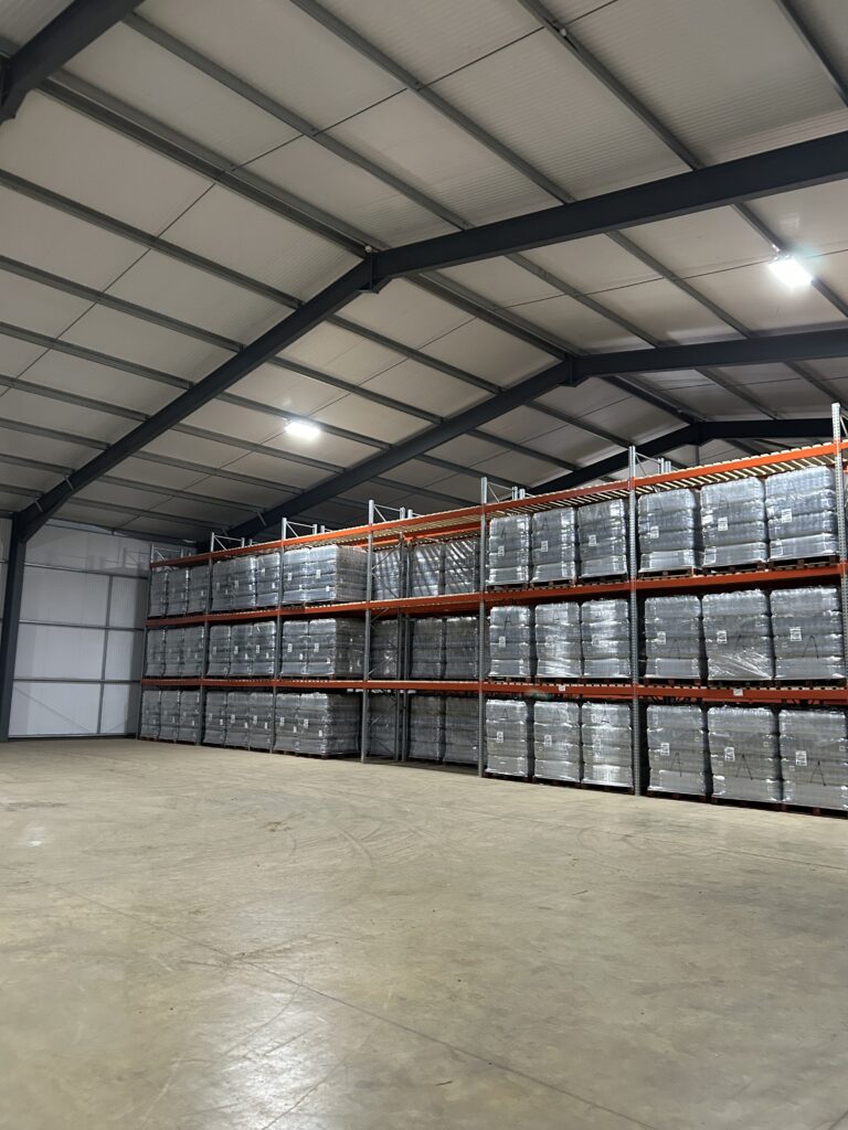 Interior of a large warehouse showing an extensive, well-organised storage area for bottled water supplies. Palletised packs of bottled water are stacked on multi-level metal racking extending across the length of the wall, with goods shrink-wrapped and uniformly arranged on wooden pallets. The storage system makes use of vertical space, with multiple tiers allowing high-volume storage while maintaining clear separation of pallets. The floor area in the foreground is wide, clean, and unobstructed, enabling safe access for handling equipment and inspection activities. The warehouse structure includes a high insulated roof with exposed steel beams and overhead lighting, providing consistent illumination. The bottled water is stored off the floor, fully wrapped, and appears protected from contamination. The consistent stacking pattern and visible labelling suggest controlled stock management, likely supporting stock rotation and traceability. The overall layout indicates a dedicated and well-maintained environment suitable for the storage and distribution of emergency drinking water supplies, with clear access routes and minimal clutter reducing operational and hygiene risks.