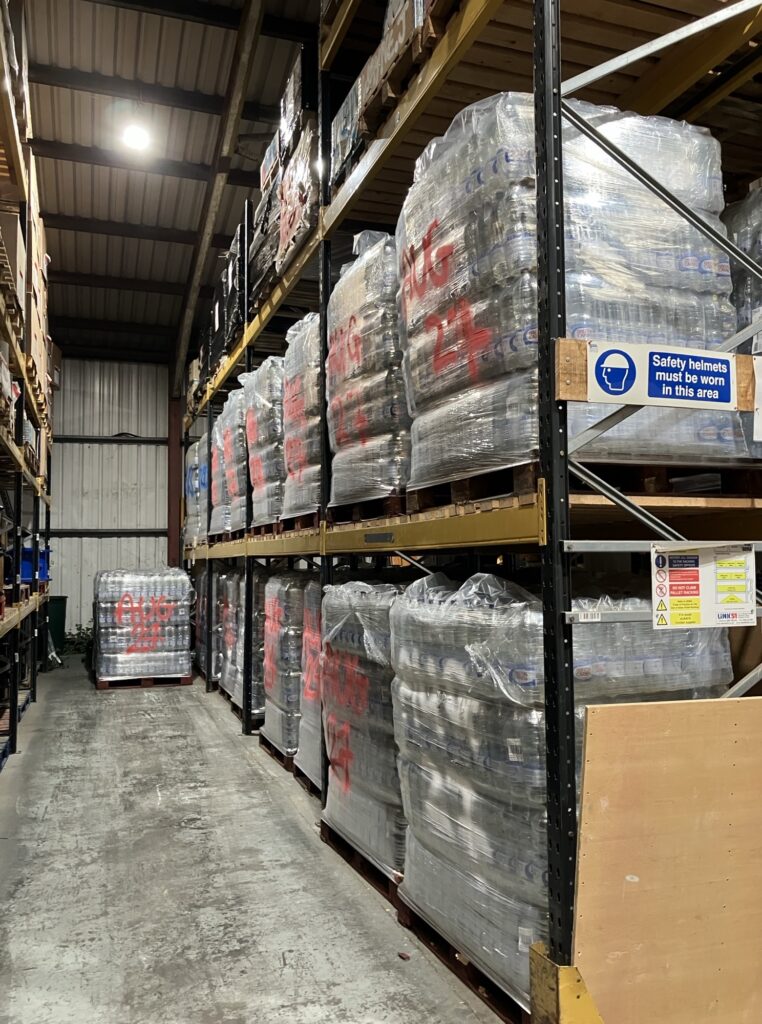 Interior of a warehouse aisle used for the storage of bottled water supplies, with palletised stock arranged on multi-level metal racking. Packs of bottled water are shrink-wrapped and stacked on wooden pallets, with several pallets marked in red spray paint with dates (for example “AUG 27”), indicating stock rotation or shelf-life management. The pallets are stored both at ground level and on upper racking tiers, maximising vertical storage space. The aisle is narrow but clear, allowing access for handling equipment. The storage environment includes overhead lighting, metal wall cladding, and exposed structural beams. Safety signage is fixed to the racking, including a sign stating “Safety helmets must be worn in this area”, and additional warning notices relating to pallet racking use. The bottled water appears to be kept elevated from the floor and fully wrapped, providing protection from contamination. The presence of visible date markings suggests an organised system for stock control and rotation, supporting the management of emergency drinking water supplies within a controlled warehouse setting.