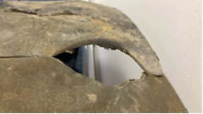 Close-up of a damaged section of a curved, weathered surface, possibly concrete or metal, with a large irregular hole exposing the hollow interior. The edges of the hole are rough and chipped, and part of a lighter surface behind the opening is visible. The outer surface shows dirt, wear, and discolouration.