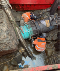 A close-up of exposed pipework in a muddy excavation, showing a large flanged joint on a water main. Water is visibly leaking from the connection and flowing into the surrounding trench. Tools and fittings are positioned nearby, indicating active repair work on a burst main.