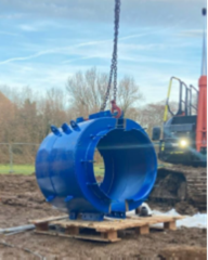 Large blue cylindrical pipe section being lifted by a chain from a crane, suspended above muddy ground and resting partly on wooden pallets. The pipe has multiple flanged openings and protruding fittings. In the background, there is construction equipment, a metal fence, and leafless trees under a clear sky.
