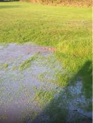 A grassy field with an area of standing water spreading across the ground, forming a shallow flood over the grass. The extent of pooling and saturated soil is consistent with flooding caused by a burst water main, with water visibly encroaching into surrounding areas.