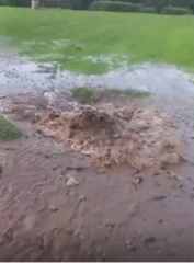A section of saturated ground where brown, sediment-laden water is forcefully erupting from the surface, creating a turbulent flow and pooling across the surrounding grass. The intensity and volume of the discharge are consistent with a burst water main, with significant disturbance of soil and localised flooding evident at the source point.