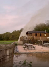 A water pipe discharging a strong stream of water across an outdoor seating area beside a small wooden building. The water sprays over tables and benches, creating wet surfaces and puddles on the ground. A wooden fence, grassy field, and trees are visible in the background under an overcast sky.