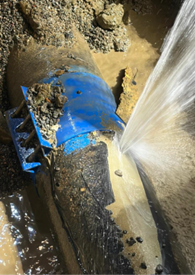 Damaged blue pipeline in a muddy excavation, with a visible break or opening in the pipe wall. Water is forcefully spraying out through the damaged section, spraying to the right. Soil, debris, and standing water surround the pipe.