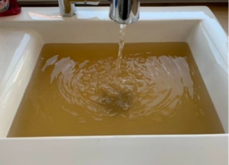 A white sink being filled from a running tap with brown, discoloured water. The water collects in the basin, showing a strong yellow-brown tint and visible disturbance where the stream enters the surface.