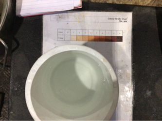 A white bucket filled with slightly cloudy water placed on a flat surface next to a printed colour grading chart. The chart shows a sequence of shaded colour blocks ranging from pale to dark. The water appears to be undergoing colour comparison as part of a flushing operation, with the bucket positioned directly over the chart for assessment.