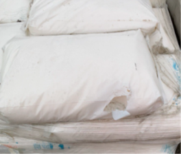 A large white bulk bag or sack placed on a pallet, with visible damage including a tear and staining along one side. The fabric appears worn and discoloured, and the bag is partially surrounded by plastic wrapping.
