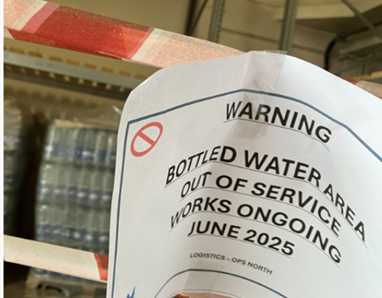 Printed warning notice attached to shelving in a storage area, stating that bottled water is out of service and works are ongoing, dated June 2025. Packs of bottled water are visible in the background behind the notice.