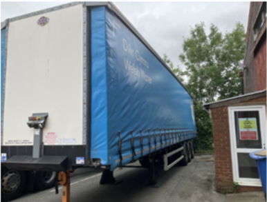 Large articulated lorry with a blue curtain-sided trailer parked in a service yard beside a brick building. A sign with red and yellow notices is displayed on a door to the right of the vehicle, and trees are visible in the background.