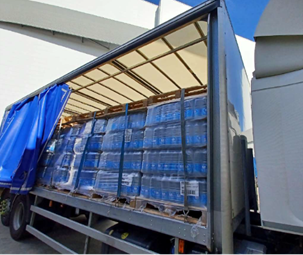 Curtain side of a lorry trailer opened to reveal pallets of bottled water stacked in rows and secured for transport. The blue curtain is partially pulled back, exposing the load inside a yard area next to a building.
