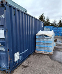 Blue shipping container positioned on a yard surface beside a stack of shrink-wrapped pallets of bottled water. The pallets are partially covered with protective wrapping, and additional blue containers are visible in the background along with trees and overcast sky conditions.