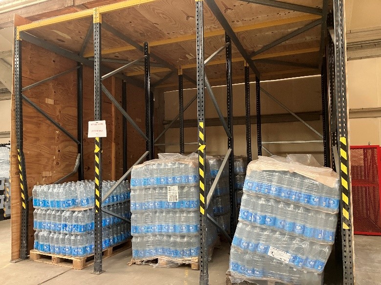 Indoor storage area with metal racking containing pallets of bottled water. Shrink-wrapped multi-packs are stacked on wooden pallets, some leaning slightly within the bays. The racking frames have yellow and black hazard markings, and a small sign is attached to one upright