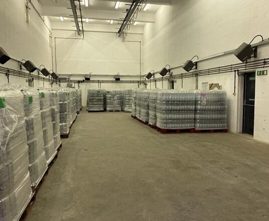 Large indoor warehouse space with multiple pallets of bottled water arranged in rows on either side of a central aisle. The pallets are shrink-wrapped and stacked on wooden bases, with additional stacks visible at the far end of the room. Overhead lighting and wall-mounted fixtures illuminate the area, and an exit door is visible on the right-hand wall.