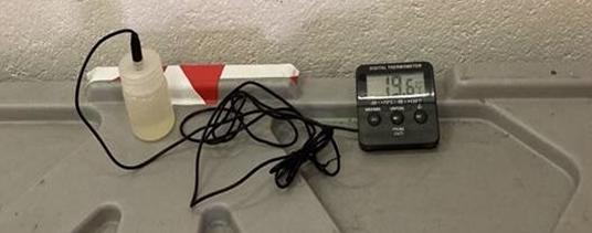 Digital thermometer probe placed on a flat surface next to a small bottle and wired sensor, displaying a temperature reading of 19.6 °C.
