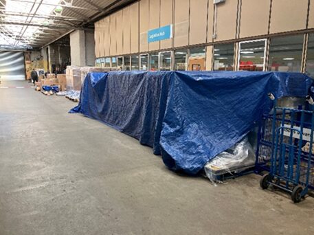 Large warehouse or depot area with pallets of bottled water lined along a wall of windows. Several pallets are covered with a blue tarpaulin, secured over stacked supplies. Additional pallets and handling equipment are visible further along the open floor space.