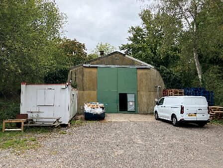 Rural yard area with a metal-clad storage building featuring large green double doors. A white van is parked to the right, and a white container unit is positioned to the left. Pallets, a skip and stacked materials are visible around the site, with trees and vegetation surrounding the area in cloudy conditions.