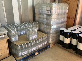 Indoor storage area showing pallets of bottled water stacked on wooden bases, with shrink-wrapped multi-packs arranged in layers. To the right, a group of white and black cylindrical containers is lined up, and to the left a small desk and drawers are positioned against the wall.