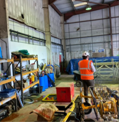Interior of a warehouse or workshop space with high ceilings and exposed metal roof beams. Shelving units on the left contain tools and equipment, while pallets covered with a blue tarpaulin are stacked towards the back. In the centre foreground, a person wearing a high-visibility orange vest and white hard hat stands facing away, near machinery and equipment including a trolley and a red container