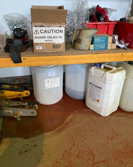 Shelving unit in a storage area containing equipment and containers. A cardboard box on the upper shelf is labelled ‘CAUTION SHARP OBJECTS INSIDE’. Below, several plastic containers and tools are stored, with visible liquid staining and pooling on the floor in front of the shelving.