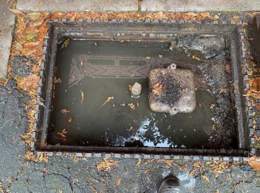 Rectangular underground chamber filled with standing water, debris, and leaf litter. A partially submerged access cover and fittings are visible within the chamber, with water reaching near the top of the structure.